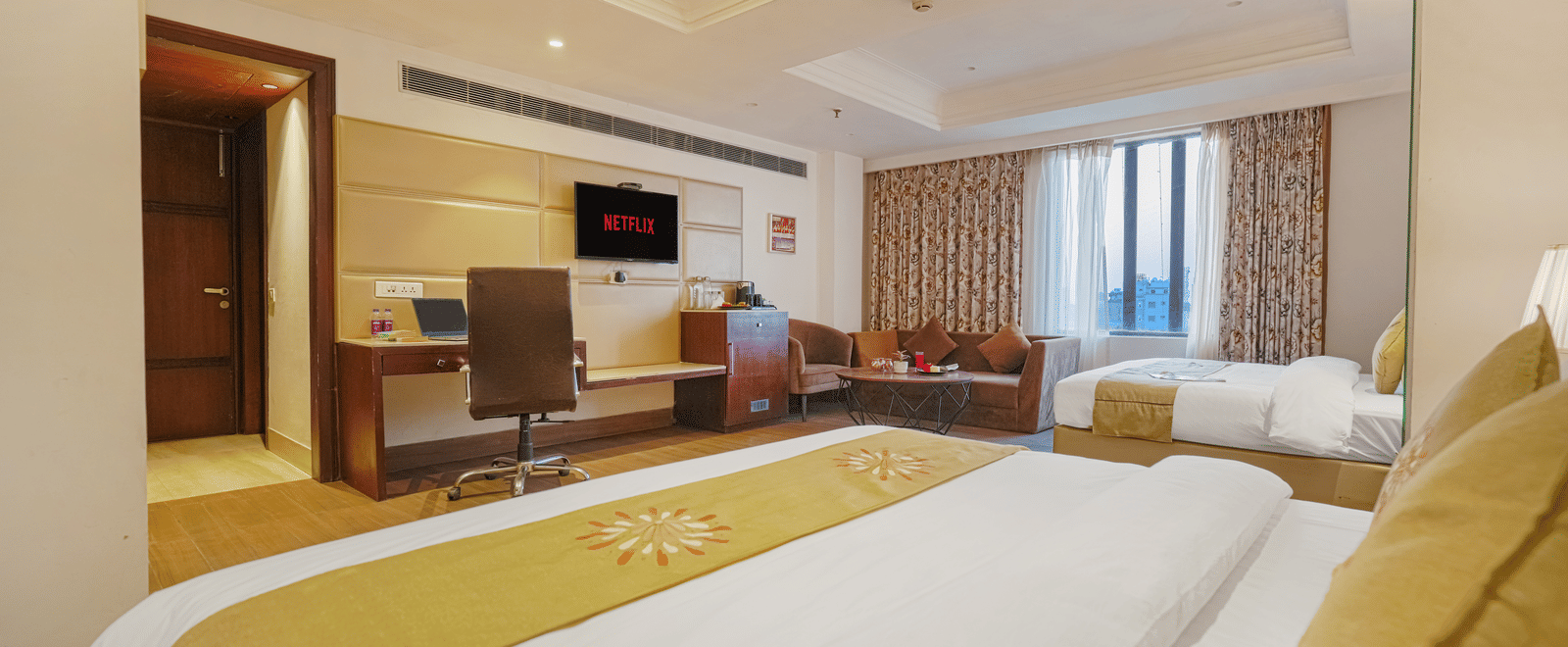 A spacious hotel room showing 2 beds with gold runners, a work desk, seating area, and wall-mounted television at BluSalzz Select City Centre, Amritsar.