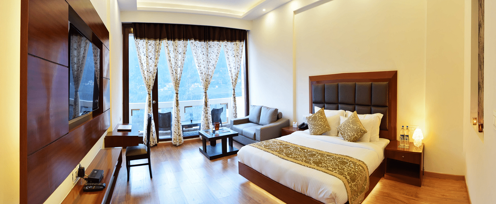 The image shows a spacious and luxurious hotel room with a king size bed, a sitting area with a sofa and coffee table, and a large window with a view.  | DLS Serenity Resort and Spa