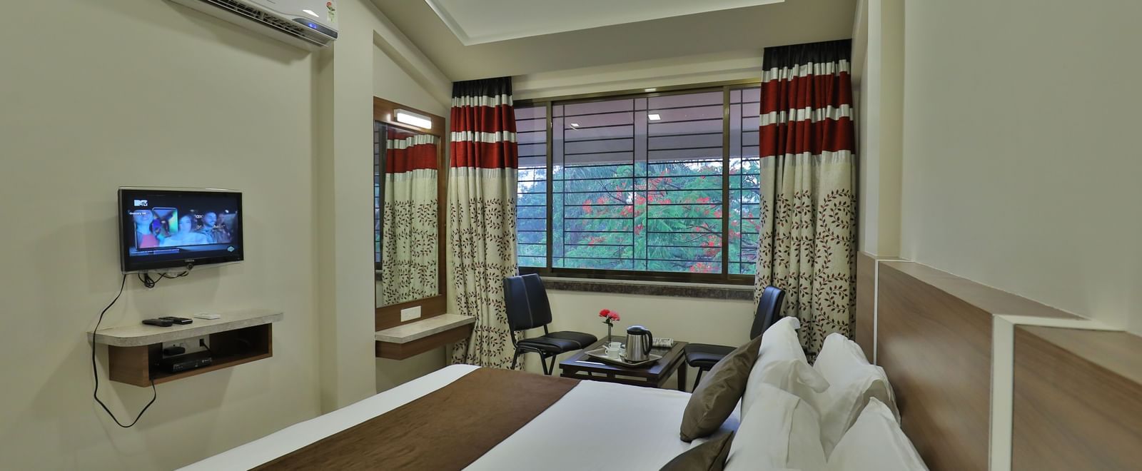 Extended view of the queen size bed facing the wall-mounted TV in the Deluxe Room of Highland Resort, that also features drawn curtains offering views of the trees outside.