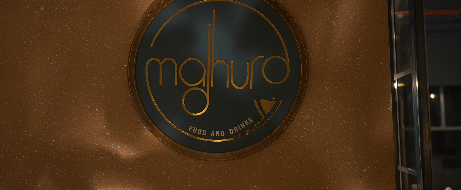 A close-up of the Madhura logo displayed on a polished brown surface at Hotel Sonar Bangla Mayapur.