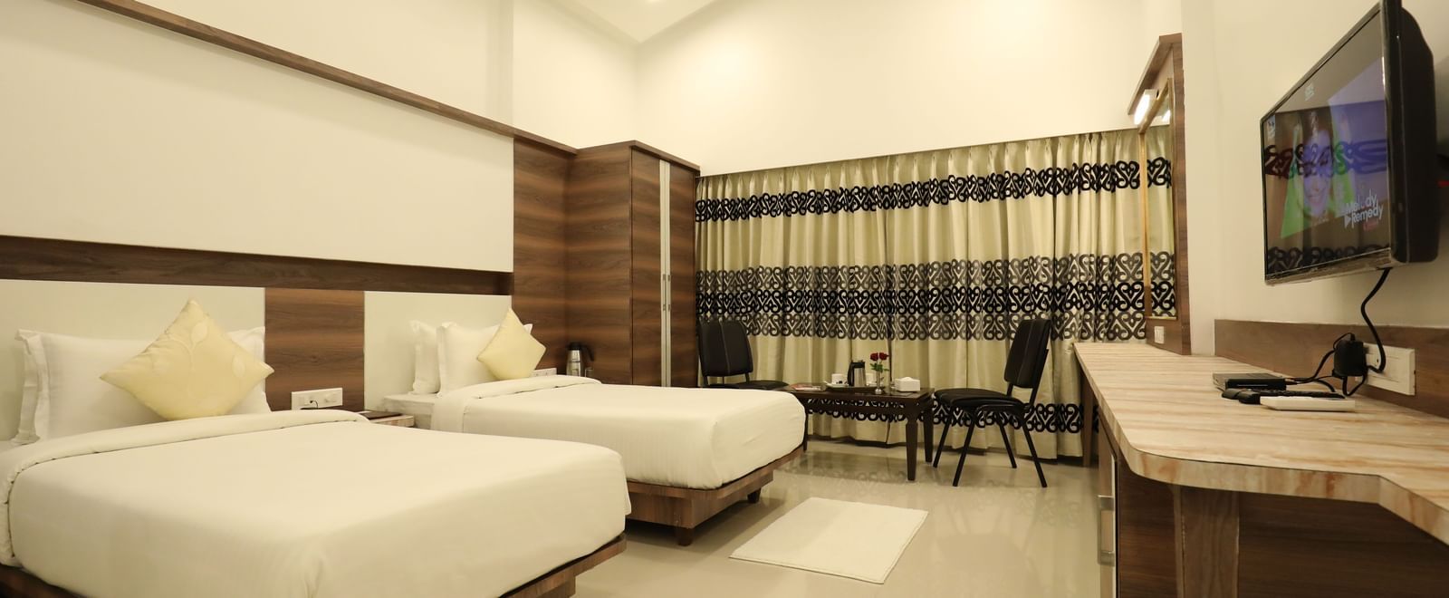 A room in Imperial Grande with twin beds, featuring a wall mounted television, and a large window with its drapes closed.