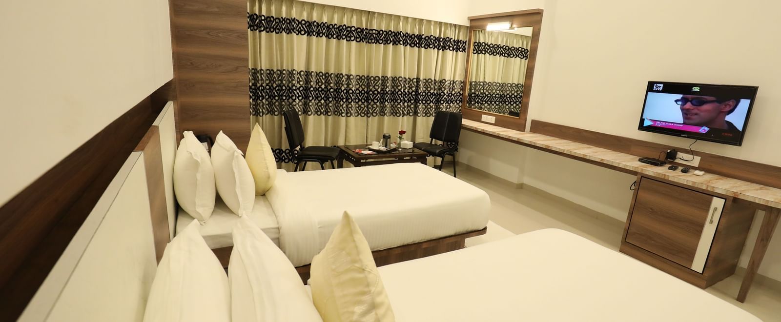 A well-lit room in Imperial Grande, featuring twin beds with pillows stacked on each, facing a wall mounted TV.