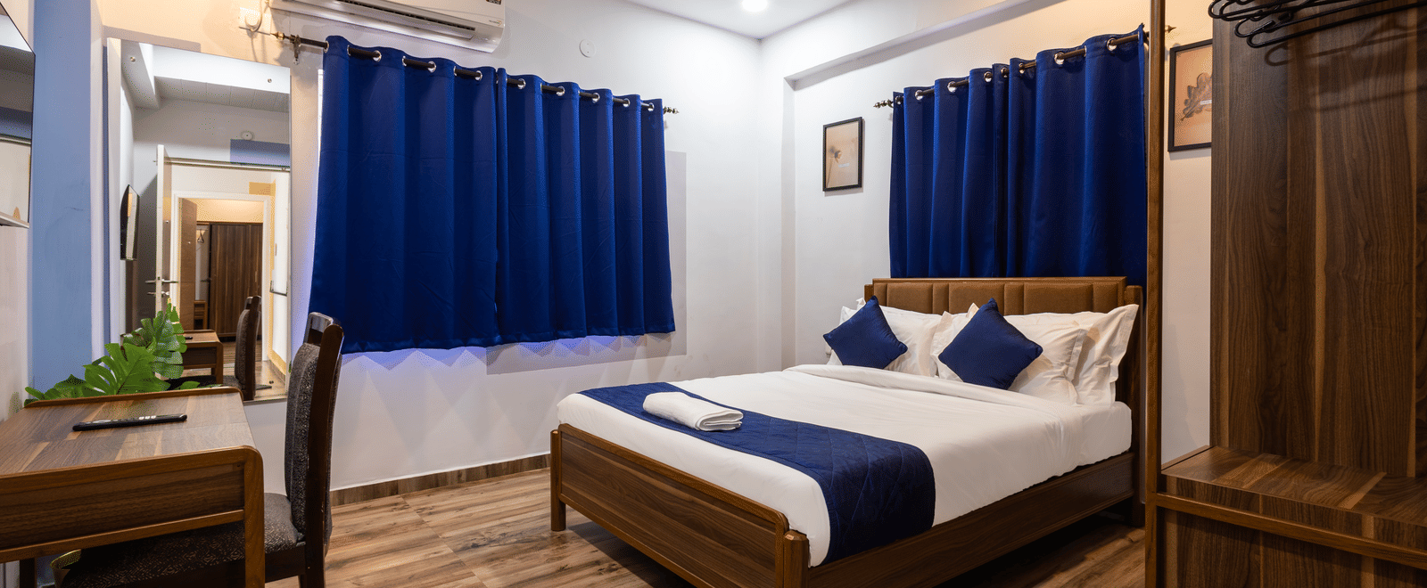 Corner view of a bedroom in the 4BHK of Kove Smart Stays, highlighting its centrally placed queen size bed, wooden wardrobe, wooden flooring, and work desk.