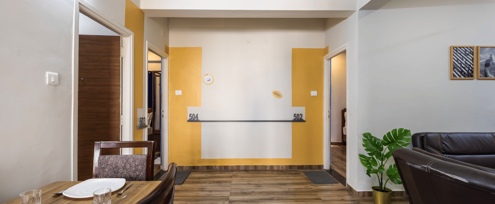 The common area of 4BHK of Kove Smart Stays, featuring wooden flooring, a wooden dining table with chairs, sofa, and 2 doorways leading to rooms inside the apartment.