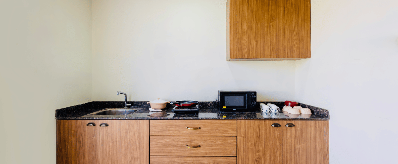Kitchen set up in one of the 4BHKs at Kove Smart Stays, seen with an oven, wooden flooring, a sink, drawers, wall-mounted shelves, and crockery placed on the countertop.