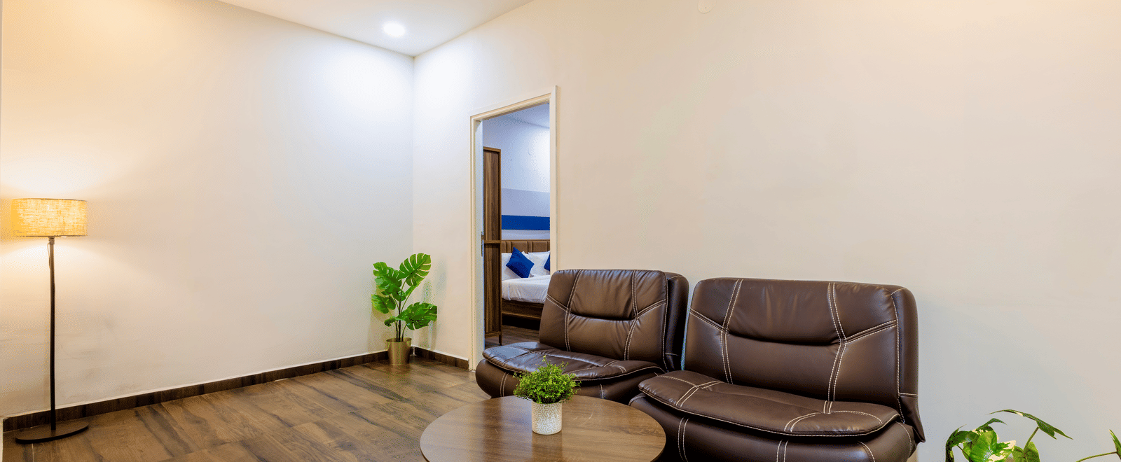 Spacious and separate living room in the Studio Room of Kove Smart Stays furnished with a multi-seater sofa with centre table, potted plants, and a lamp.