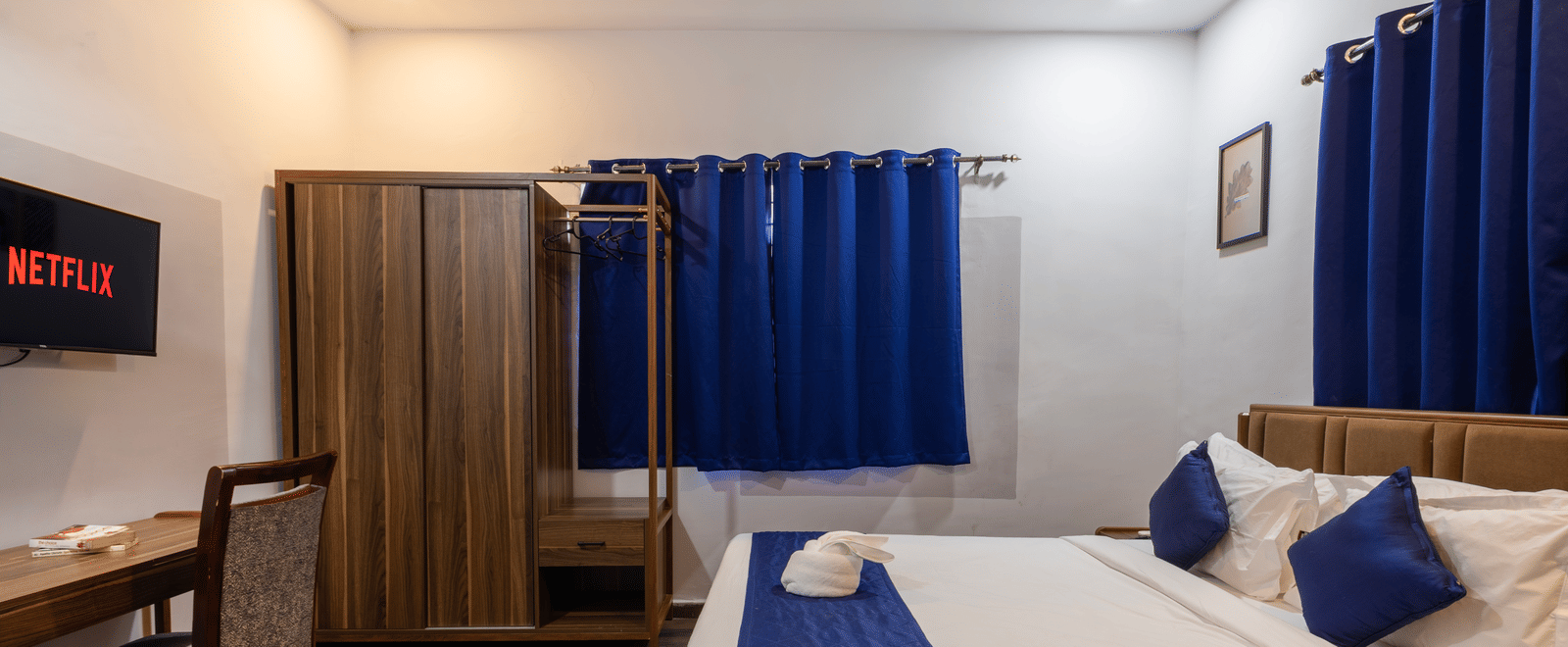 Side view of the Deluxe Room featuring the queen size bed, wooden wardrobe, wooden flooring, closed curtains, wall-mounted TV and work desk.