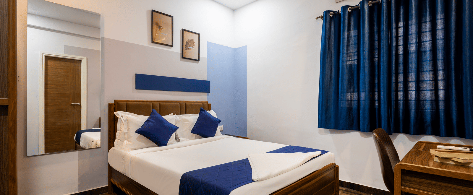 Wide view of the bedroom of the Studio Room showcasing the queen size bed, wall-mounted mirror, closed curtains, and work desk.