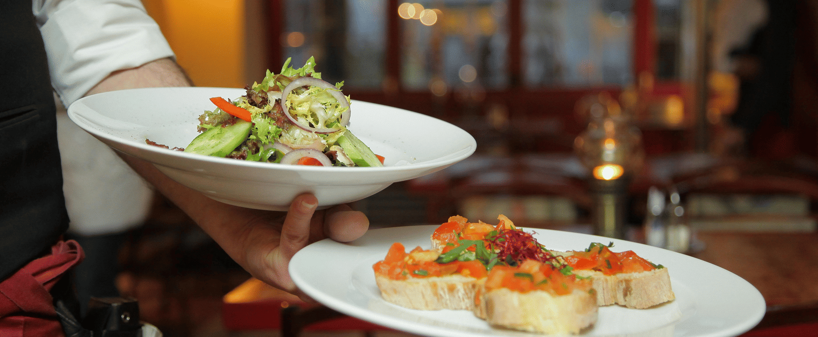 A person in a restaurant holding two plates with delicious dishes in it that are well plated.