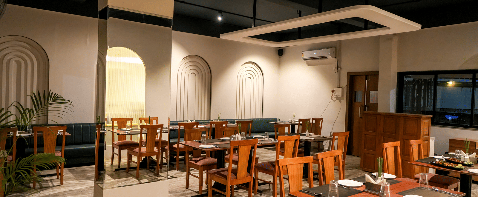  An interior view of the bright dining hall with white walls, wooden chairs, and recessed archways at Pravasa Hotel, Kolhapur.