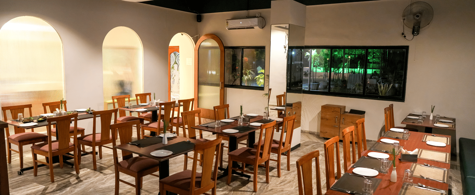 A well-appointed indoor dining room with wooden tables and chairs, white walls, and elegant archways at Pravasa Hotel, Kolhapur.