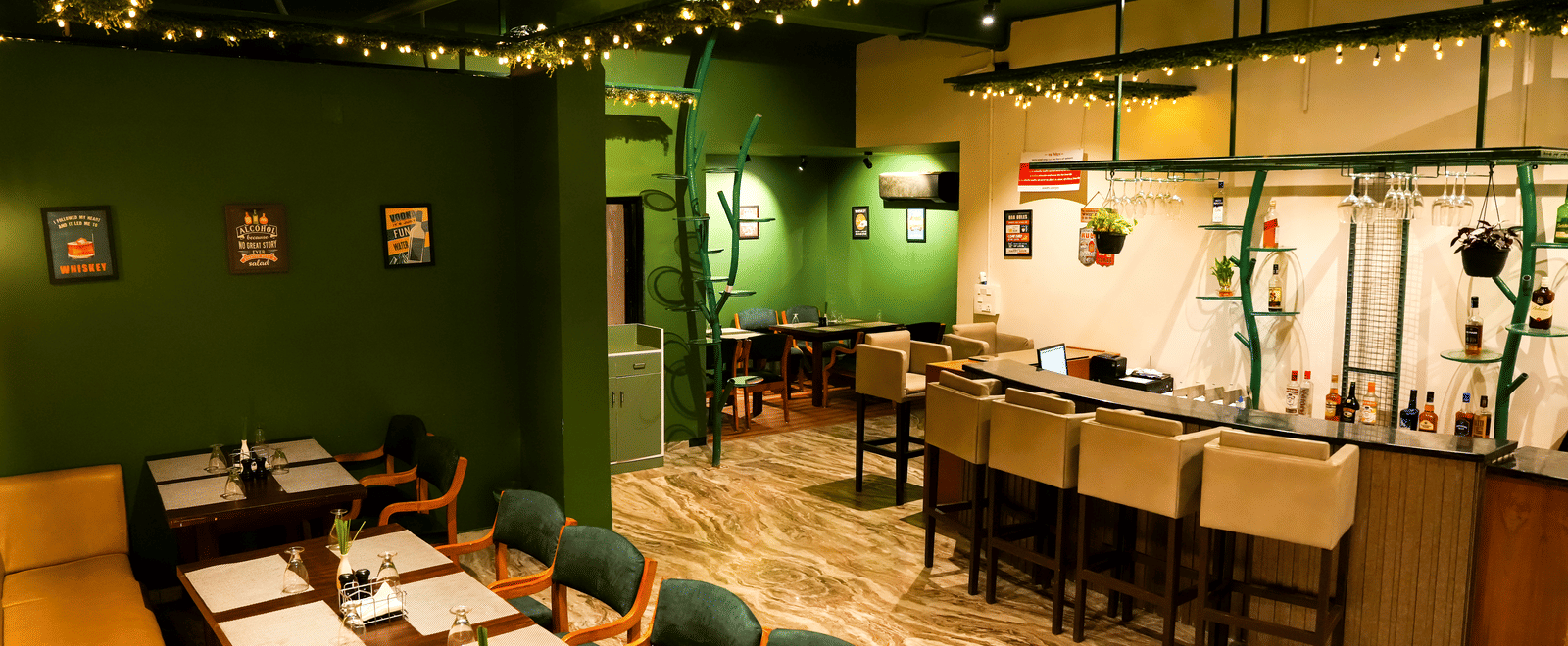  The interior of a casual dining room at Pravasa Hotel, Kolhapur, featuring green walls, simple laid tables, and a breakfast bar area.