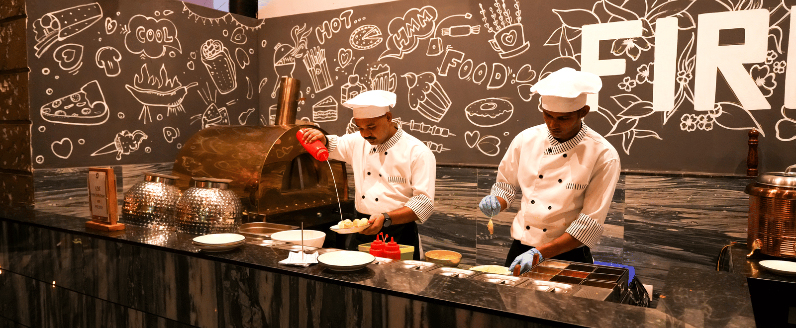  Two chefs preparing food at a live cooking station with a pizza oven and a chalkboard wall in Pravasa Hotel, Kolhapur.