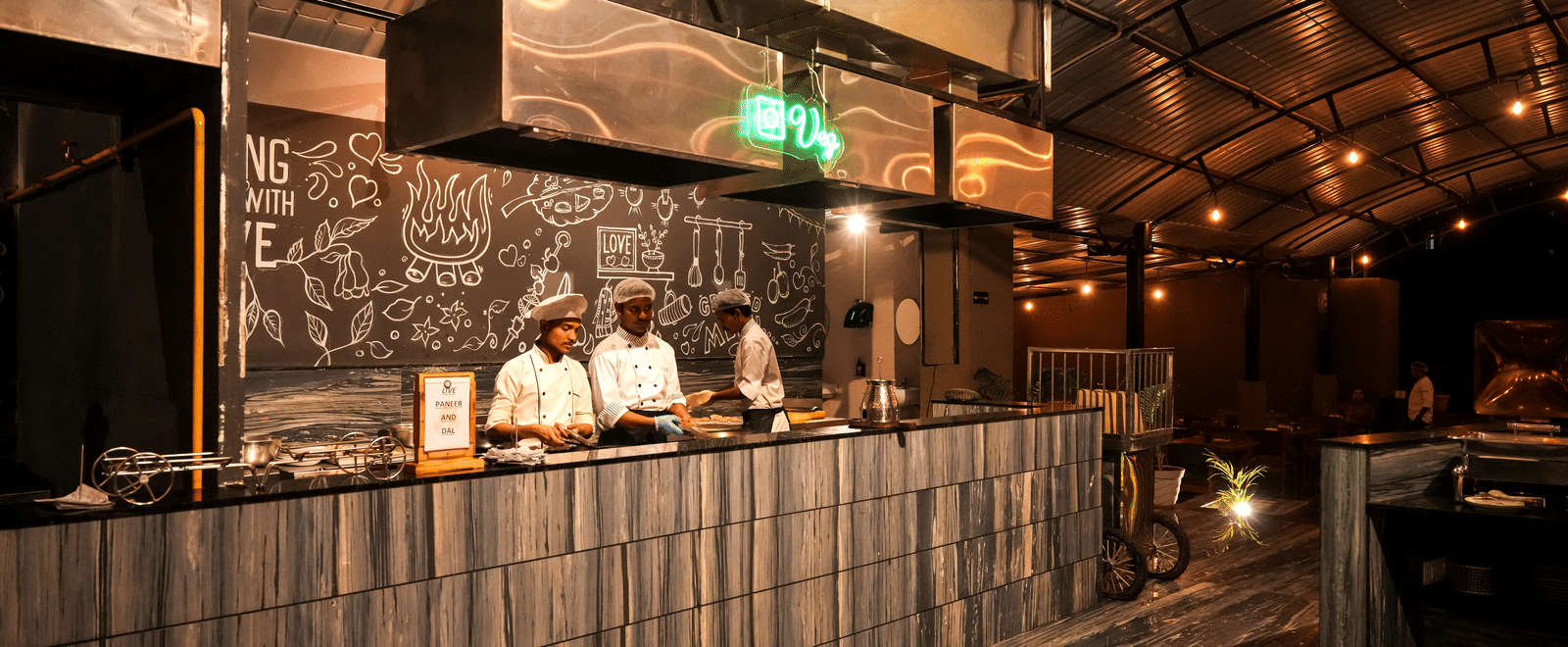  Chefs are busy working behind a modern, textured counter in the open kitchen area of Pravasa Hotel, Kolhapur.