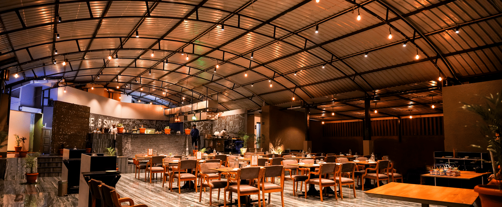  A wide view of the spacious dining hall with wooden tables and chairs under a curved, industrial-style roof at Pravasa Hotel, Kolhapur.