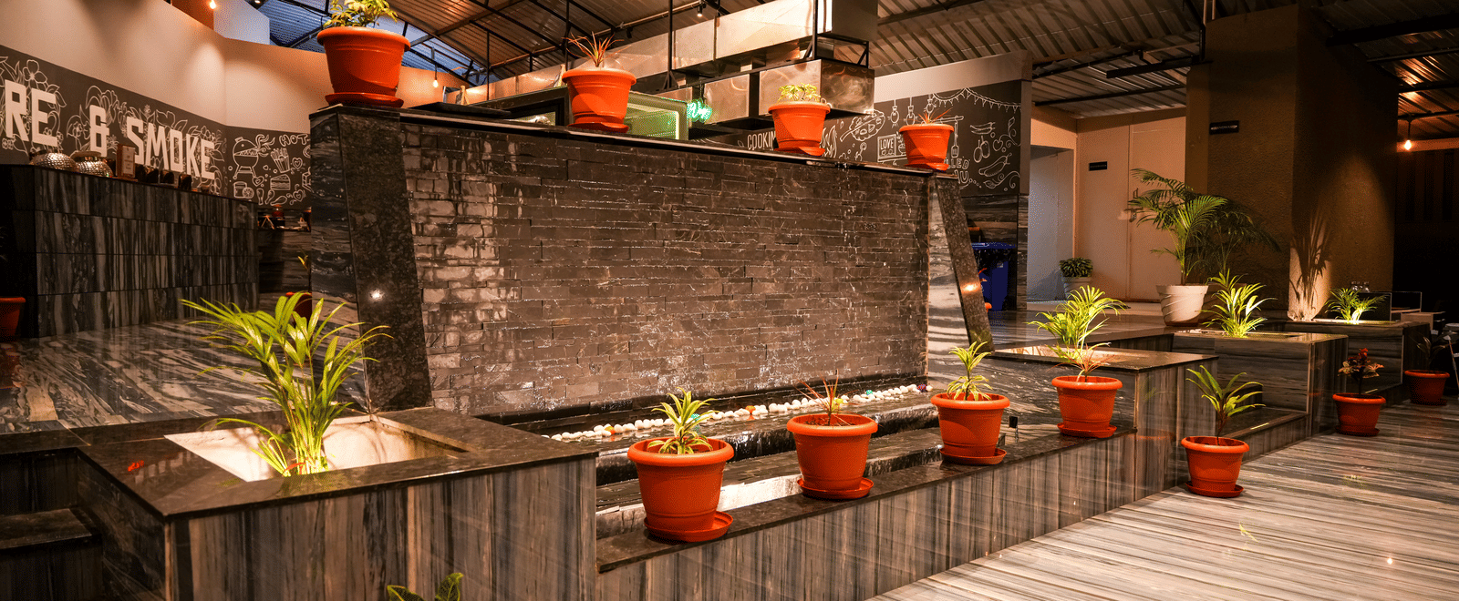 A stylish indoor seating area with plants in terracotta pots and a corrugated iron roof at Pravasa Hotel, Kolhapur. 