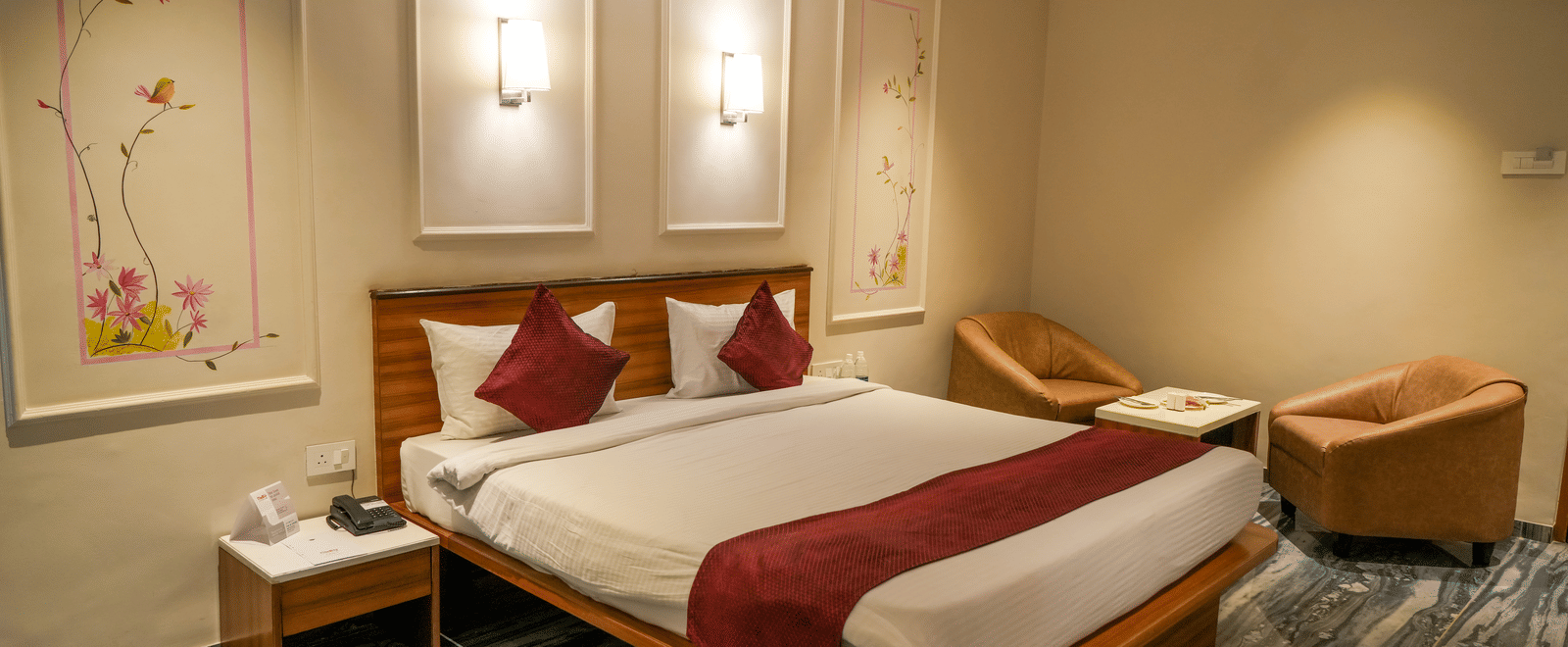 A stylish deluxe room featuring a double bed, two comfortable armchairs, and nature-inspired wall art at Pravasa Hotel, Kolhapur.