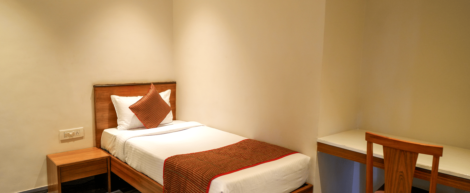 A compact and neat single bedroom with a wooden bed, a study desk, and marbled-effect flooring at Pravasa Hotel, Kolhapur.