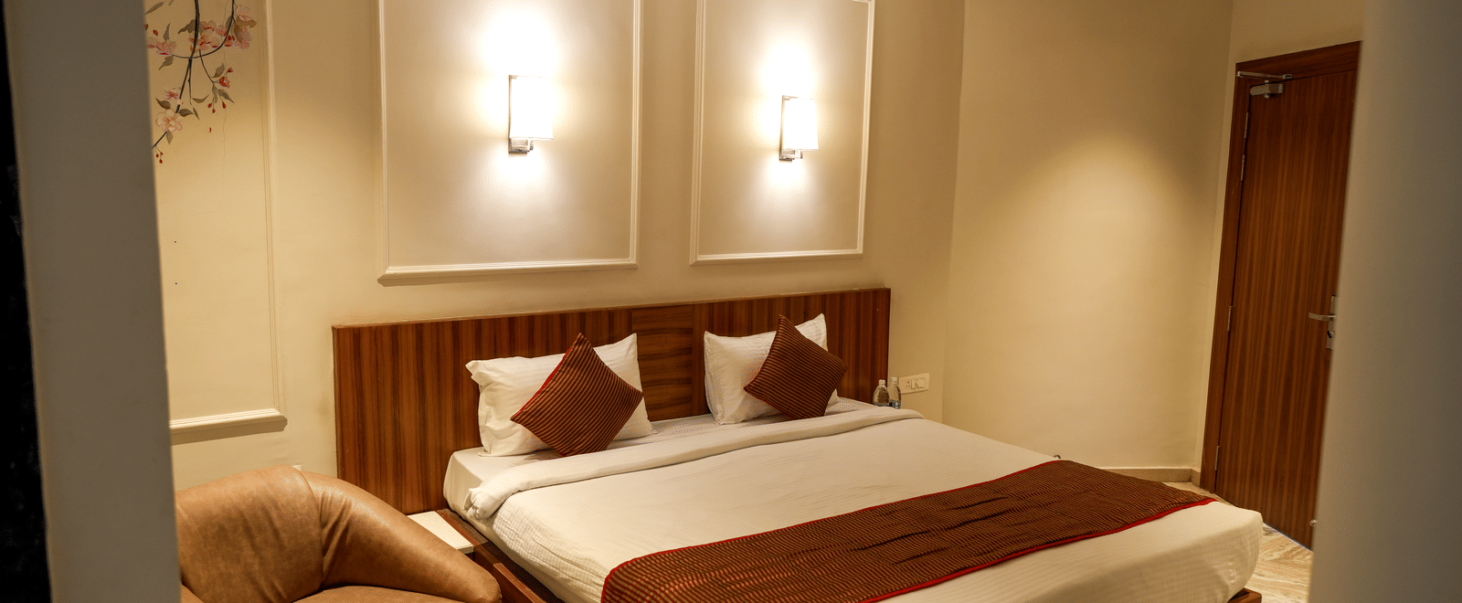  A modern guest bedroom at Pravasa Hotel, Kolhapur, featuring a double bed, subtle wall panel moulding, and a tan armchair. 