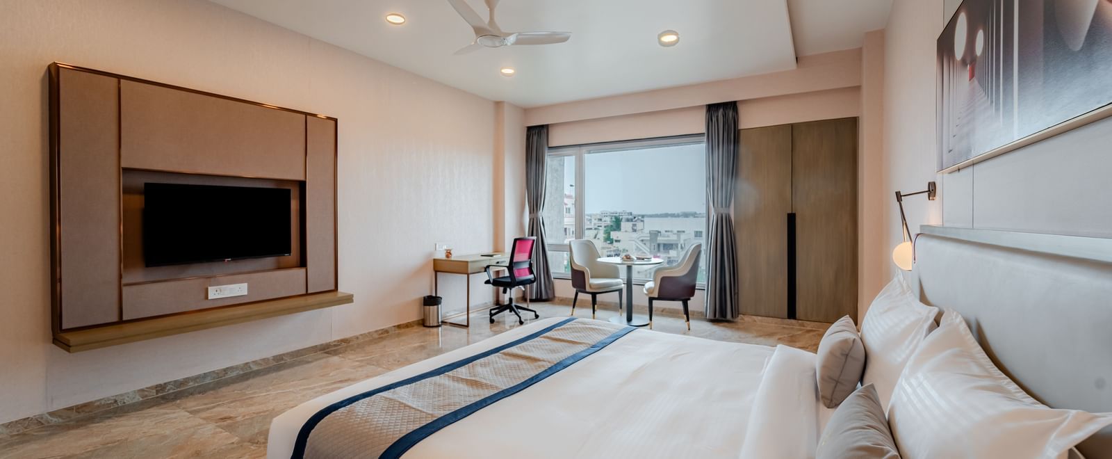  A wide angle shot of a hotel bedroom with a large bed, TV, and a desk, well-lit | Premium Room | Pravasa Hotel, Vijayapura