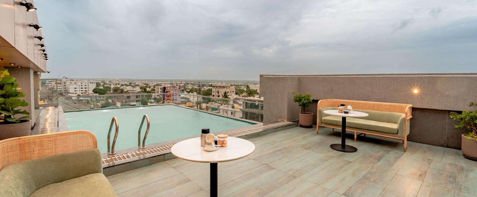 The rooftop swimming pool with clear water, surrounded by a tiled deck and some modern outdoor seating, offering a panoramic view of a city under a cloudy sky | Pravasa Hotel Vijayapura