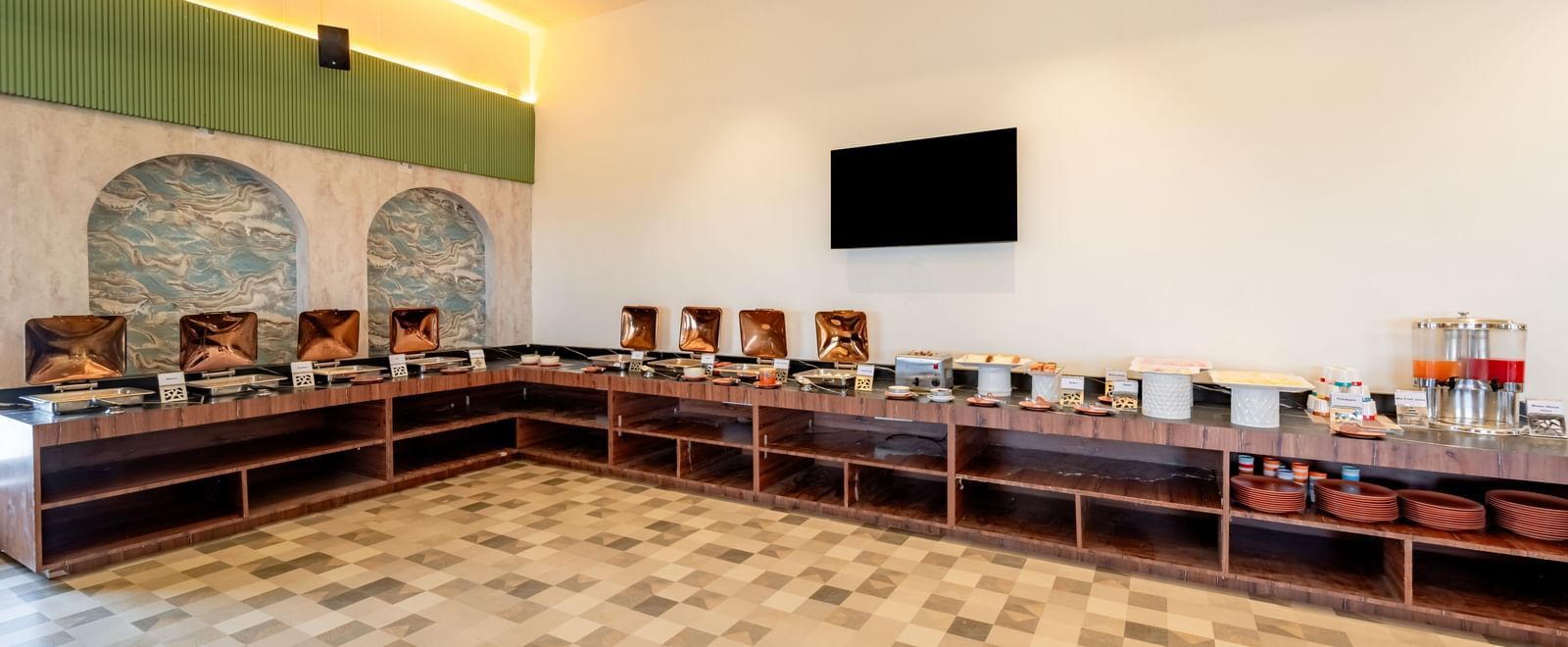 Long buffet counter with an assortment of breakfast items and a TV screen | Pravasa Hotel, Vijayapura
