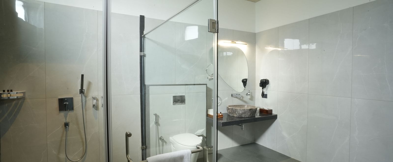 A bathroom of Pool View Room equipped with a glass-enclosed shower, a toilet, and a sink, featuring clean lines and a minimalist design at The Qualia Resort Club & Brewery, Udaipur.