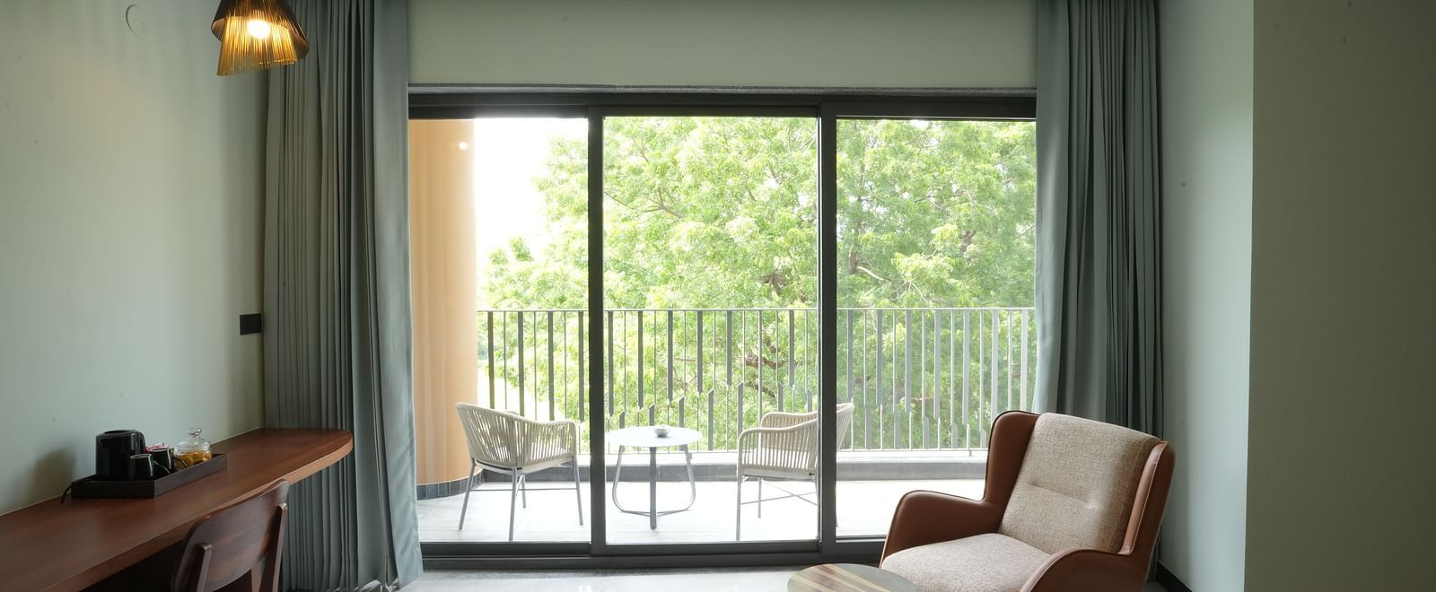 Club Suite with Valley View Sitout depicts a contemporary living space featuring a desk, chair, lounge chair, and a large window offering a view of a balcony and greenery at The Qualia Resort Club & Brewery, Udaipur.