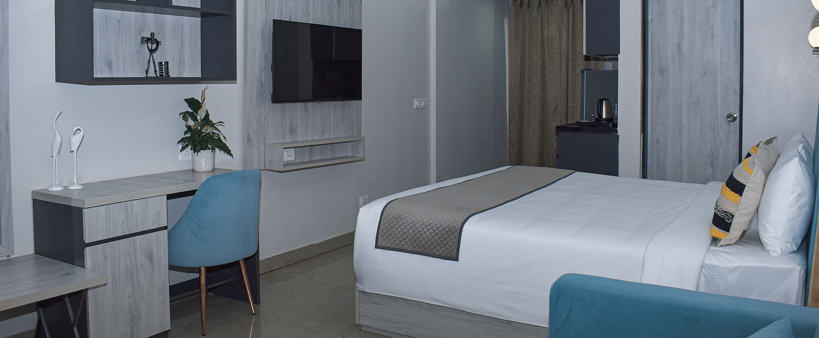 A hotel room with a bed, a blue sofa, a desk and chair, a wall-mounted TV, a kitchenette area, and a wall cabinet - Urban Suites by BluSalzz, Jaipur