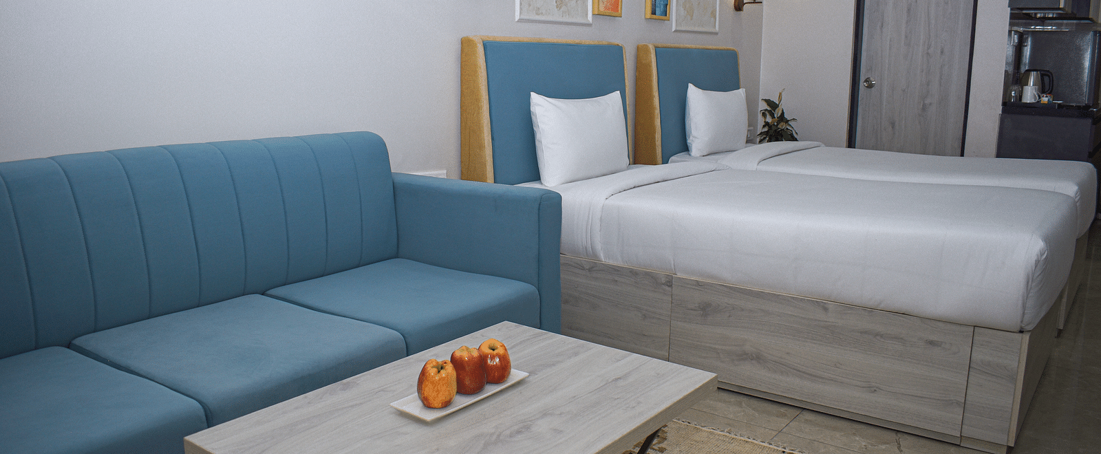 A hotel room with two twin beds, a blue sofa, a light wood coffee table with fruit, and a kitchenette area - Urban Suites by BluSalzz, Jaipur