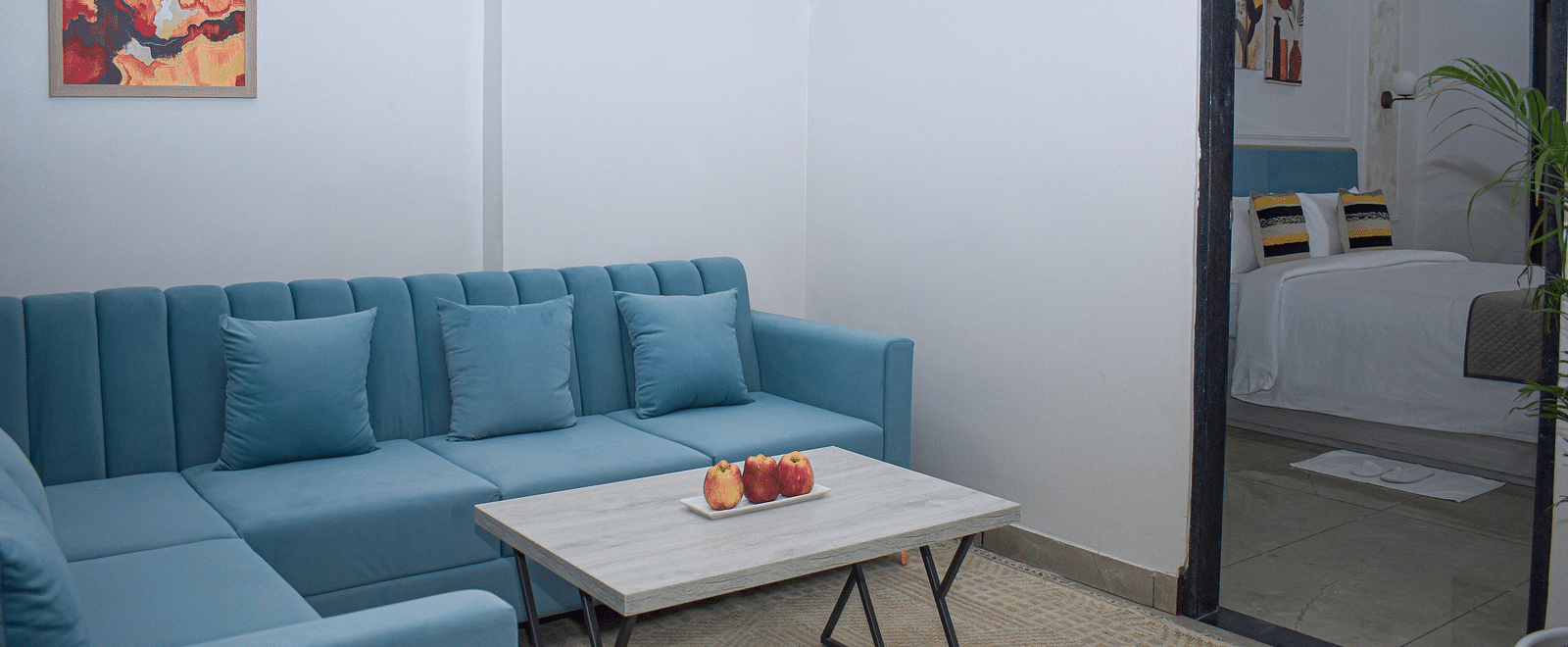A hotel room living area with a blue sofa, a light wood coffee table with fruit, a rug, and a view into the bedroom - Urban Suites by BluSalzz, Jaipur