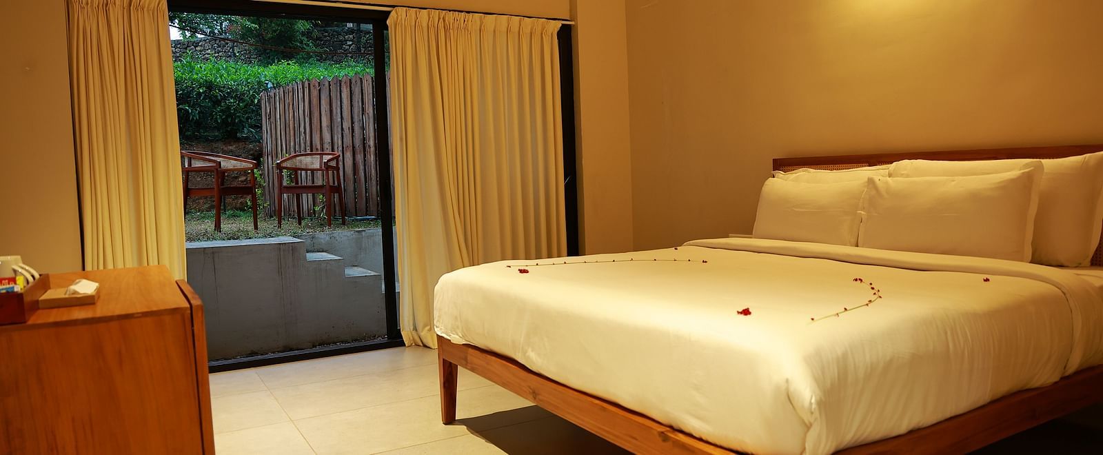  A hotel room at Vaasta Vagamon featuring a bed with subtle petal decor, a wooden cabinet, and a sliding door leading to an outdoor patio.