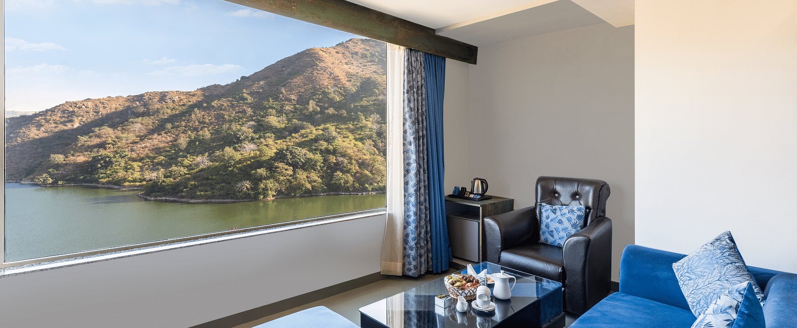 Living area with a blue couch and a large window with a view of mountain and water - Via Lakhela Resort & Spa