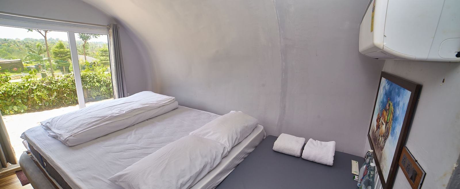 The interior of a modern, capsule-style accommodation unit featuring two single beds with white mattresses and a small bedside table, with a window offering a view of greenery. - Vintage Trails by Ilara