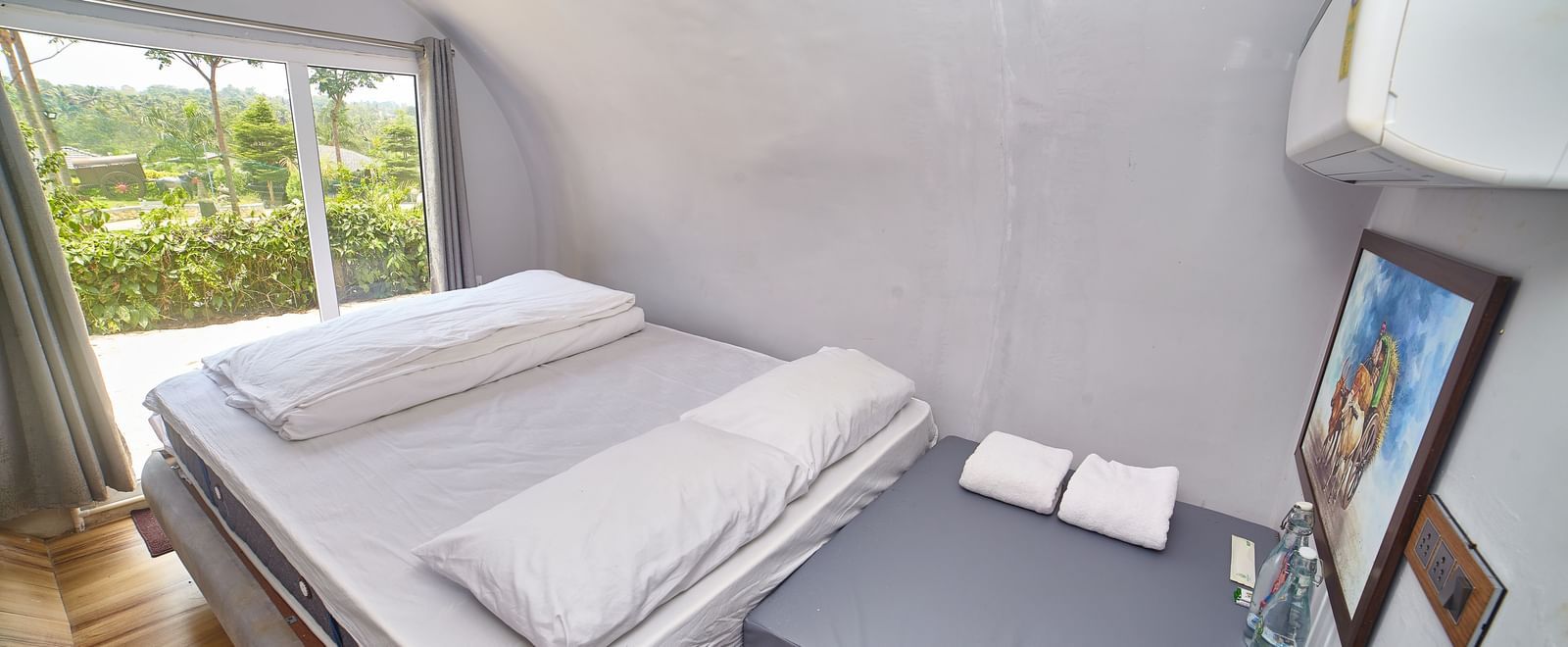 The interior of a modern, capsule-style accommodation unit featuring a single bed with a white mattress and a small bedside table, with a window offering a view of greenery. - Vintage Trails by Ilara