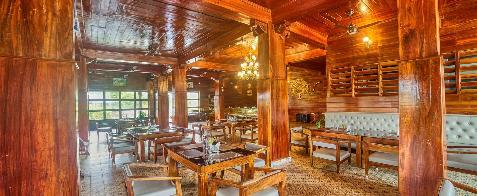 A spacious, warmly lit dining or common area with polished wooden floors, intricate wooden ceilings, and numerous tables and chairs, showing more of the window view. - Vintage Trails by Ilara