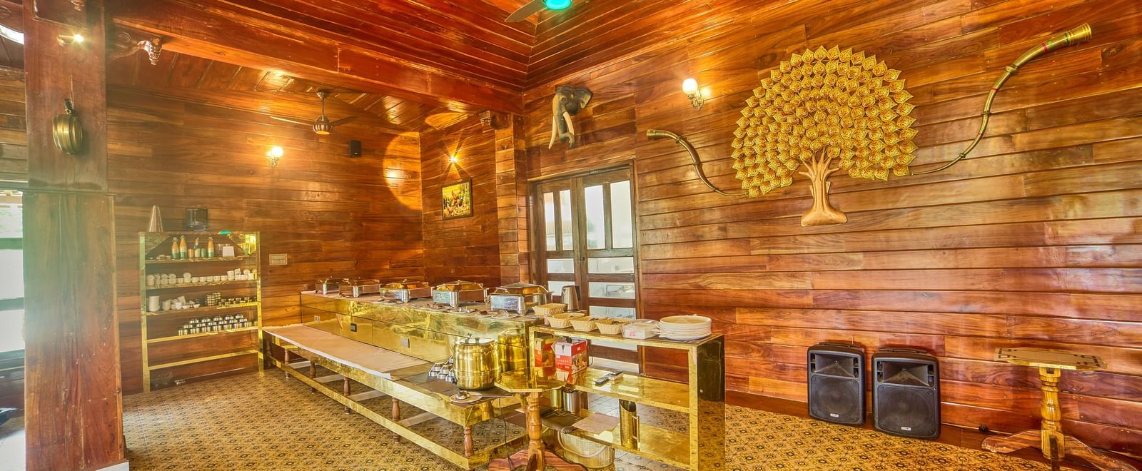 A warmly lit room with rich wooden panelled walls and ceiling, featuring decorative lighting, colourful accents, and an open seating area, possibly a dining or common space. - Vintage Trails by Ilara