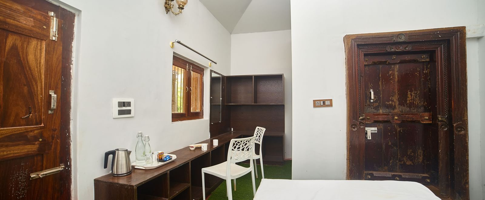 A bright hotel room featuring a bed with white linens, wooden furniture including a cabinet and a white chair, and two dark wooden doors. - Vintage Trails by Ilara
