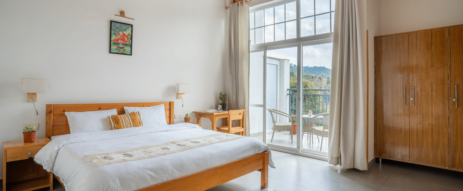 A wide shot of a Ziran Retreat bedroom with a large bed, a wooden bedside table, and large glass doors open to a balcony with a scenic view.