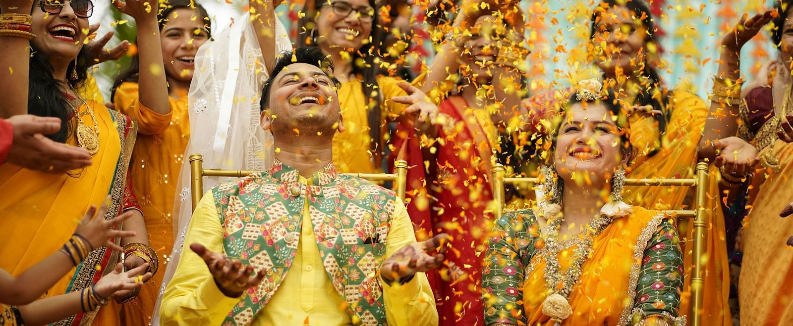 A close up shot of a couple sitting in the middle surrounded by tother people all dressed in yellow as flower petals fall over them.