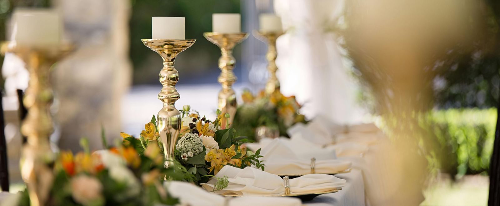 Close up shot of a dining table with golden coloured candle stands each holding a white candle and several flowers on the table.