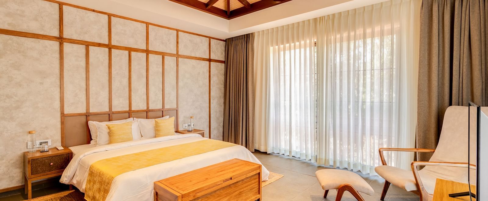 A cosy bedroom with a pitched wooden ceiling, a king-sized bed, warm tones, and soft natural light with  a large window in the background paired with semi transparent curtains  impressions of sunlight - BluSalzz Villas - Alocassia, Anjuna