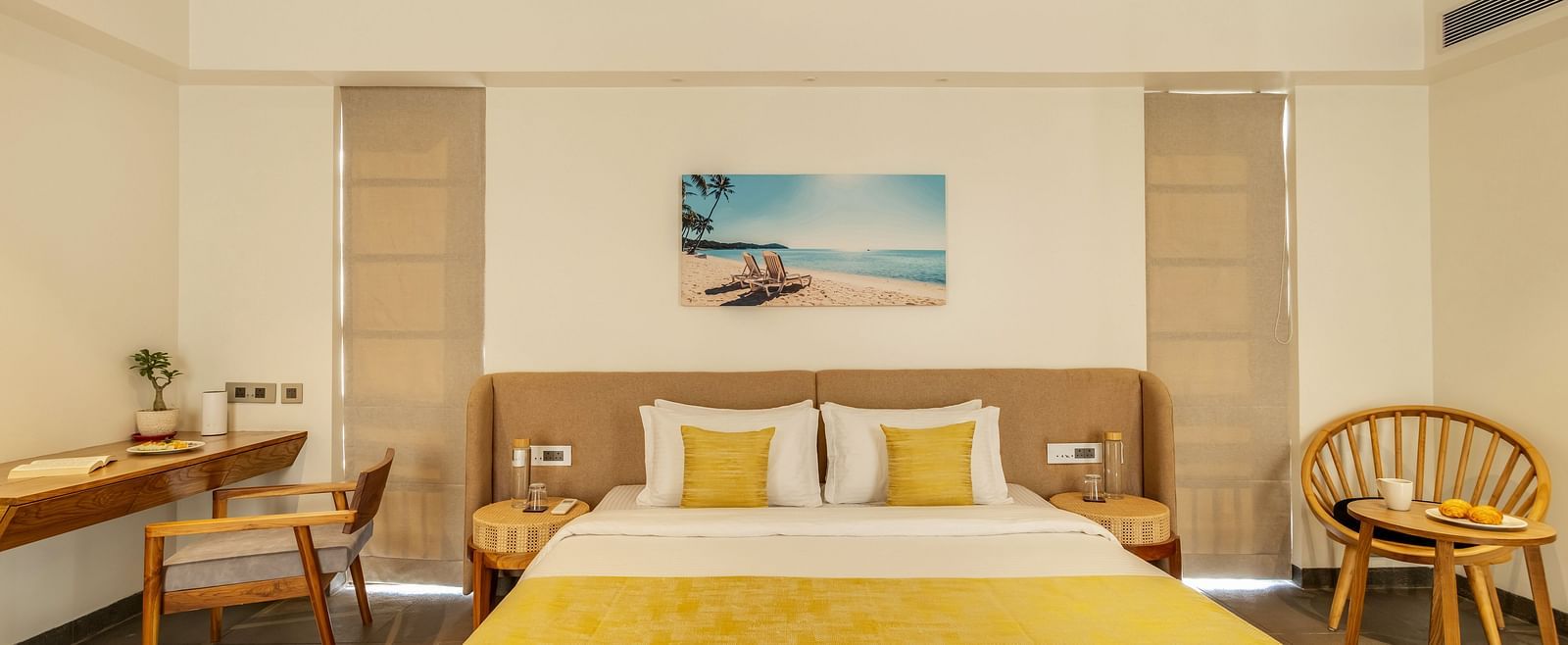A neatly designed bedroom featuring a double bed with yellow-themed decor, a writing desk, and full-length windows with flowing curtains - BluSalzz Villas - Alocassia, Anjuna
