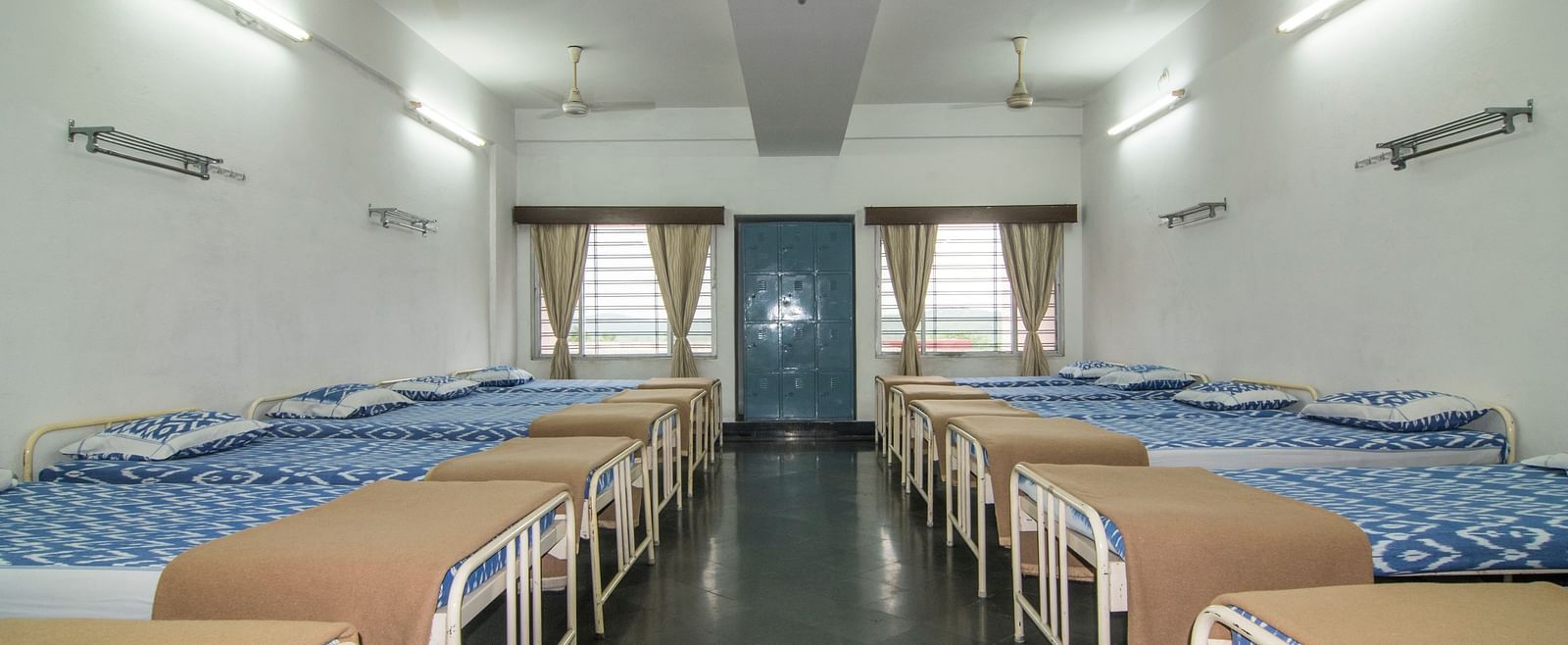 A full view of beds with bedding arranged in two rows in a room at Hotel Sahara - Spacious Dormitory in Ramoji Film City, Hyderabad.
