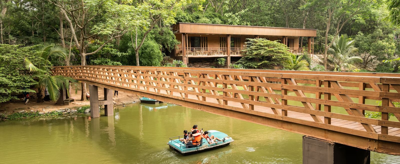 A wooden bridge spans across a calm body of water where a small boat is visible, surrounded by abundant green foliage | Lake View Restaurant
