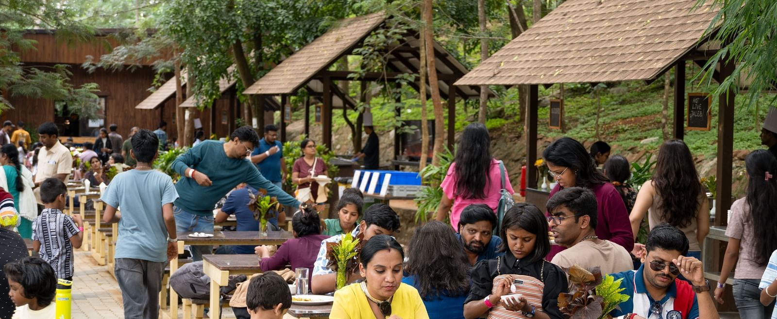 A lively outdoor dining scene shows many people gathered at tables under rustic shelters in a lush, green setting | Lake View Restaurant