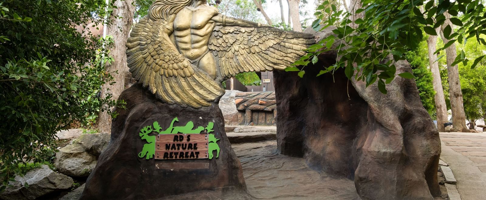 A large stone eagle sculpture stands at the entrance of a lush, green pathway leading into a natural-themed area