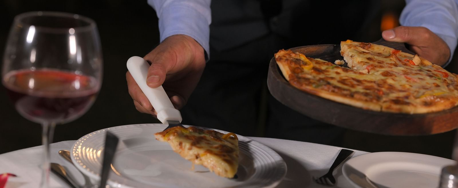 A table set for dining with cutlery, a glass of wine and a person serving a slice of pizza at Royal Comfort Regency.