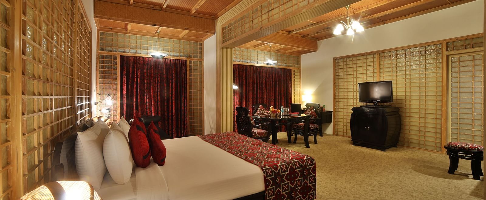 A warmly lit hotel room with traditional decor, a large bed, and wooden lattice work adding to the ambiance A grand hotel suite with an opulent red and white decor, large bed, and seating area, reflecting a traditional style at Sitara Luxury Hotel in Hyder