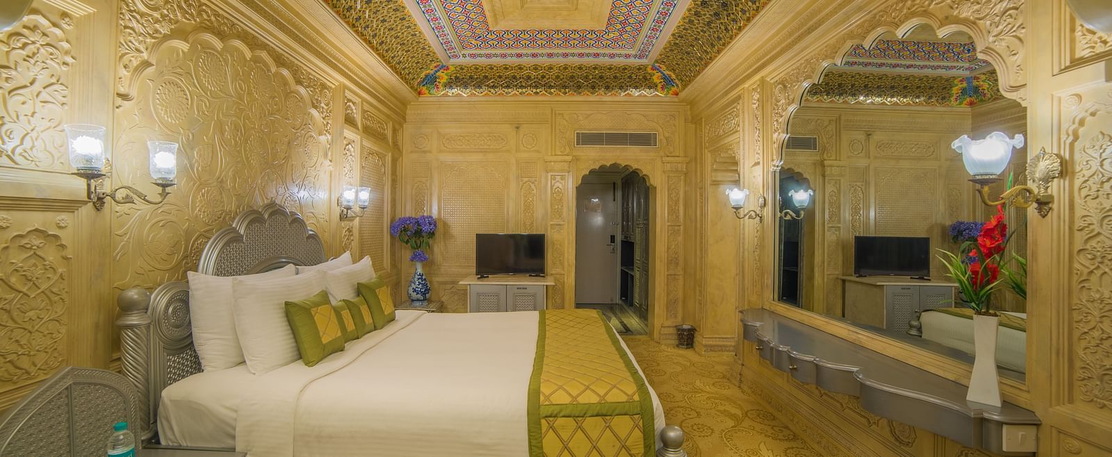 A palatial room with exquisite carvings, muted tones, and plush bedding for a regal experience A grand hotel suite with an opulent red and white decor, large bed, and seating area, reflecting a traditional style at Sitara Luxury Hotel in Hyderabad near Ram